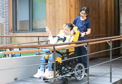 녹색 상의에 청바지를 입은 휠체어 이용자와 파란 옷을 입은 지원인이 행복마을 안에 설치된 경사로를 지나고 있는 사진, 경사로 양쪽에는 핸드레인일 설치되어 있다