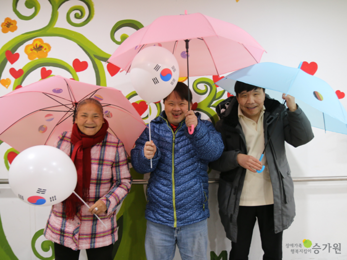 태극기가 그려진 우산과 풍선을 든 채로 활짝 미소 짓는 성인 장애가족 3명의 모습. 오른쪽 하단 장애가족 행복지킴이 승가원 CI
