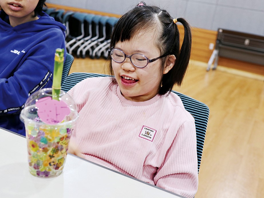 여성 장애아동이 완성된 반려식물을 바라보며 미소 짓는 모습. 양갈래로 머리를 묶고 안경을 착용하였고, 옷은 분홍색 상의를 입고 있다.