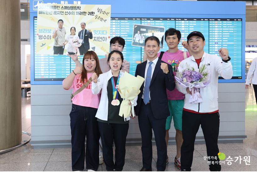 공항에서 메달 수상을 축하하는 꽃다발과 배너를 들고 있는 모습. 김용진 관장과 그 외 근무가족들이 메달을 목에 건 문지연 선수와 함께 화이팅을 외치는 모습. 오른쪽 하단 장애가족 행복지킴이 승가원CI
