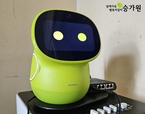 1인고령장애인을 위한 반려 로봇이 컴퓨터 본체 위에 올려져있다. 반려로봇은 연두색이며 화면에는 동그란 눈이 그려져있다. 우측상단 장애가족 행복지킴이승가원 ci삽입