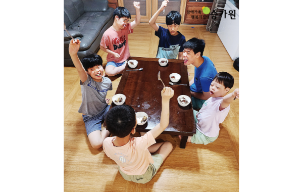 왼쪽 상단에 장애가족행복지킴이 승가원ci / 6명의 남성 장애아동들이 네모난 나무 테이블에 빙 둘러앉아 그릇에 담긴 케이크를 먹고있음. 5명의 장애아동은 포크를 든 손을 번쩍 들고 있고, 오른쪽 파란 옷을 입은 장애아동은 밝게 웃고있음.