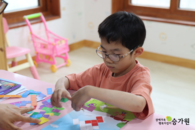 승가원행복마을 놀이치료실에서 인지치료(시지각협응)에 참여하고 있는 장애 아동. 퍼즐 혹은 테트리스 모형 같은 교구를 집어 틀에 맞추어 끼워 넣는 연습을 하고 있음. 오른쪽 하단 장애가족 행복지킴이 승가원CI