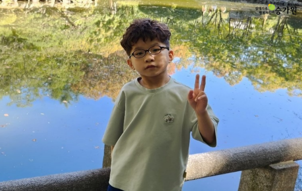 수많은 나무가 비추는 연못 돌다리 계단 위에서 짧은 파마머리에 동그란 안경을 쓴 남자아이가 손가락으로 브이 포즈를 하고 있는 사진. 오른쪽 상단에 장애가족 행복지킴이 승가원ci