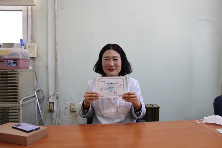 100회 나눔증서를 들고 수줍게 웃고 계시는 박지현 후원가족님 모습. 제약회사에 근무하고 계셔서 흰 가운을 입고 계시는 모습이다.