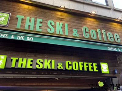 더 스키 카페의 간판의 모습. 간판은 2개로 나누어져있고 나무로 이루어져있다. 윗 간판은 초록색 영어 글씨로 'THE SKI & Coffee'라고 적혀져있다. 글씨 왼쪽 부분에는 정사각형 모양 간판에 'THE SKI'라고 적혀잇고, 스키를 타는 사람의 형상이 그려져 있다. 바로 아래에는 영어로 커피 앤 더 스키라고 써 있는 초록색 긴천이 있다. 밑 간판은 형광 연두색 글씨로 'THE SKI & COFFEE'라고 적혀 있고, 글자 양쪽에 더 스키 카페의 로고가 붙어 있다. 윗 간판에 상단에는 노란색 조명이 4개가 달려있고, 간판 위에는 회색 건물이 보인다. 우측하단 장애가족행복지킴이 승가원CI